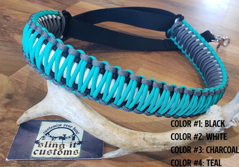 May include: A teal, white, black, and charcoal braided paracord sling. The sling is made with a woven pattern and has a black adjustable strap. The text "customize your hunt sling it customs" is on a card below the sling.