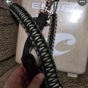May include: Black and olive green paracord shoulder strap with a metal clasp and a matching wrist strap. The strap is attached to a beige cooler. The cooler has the brand name "ENGEL".
