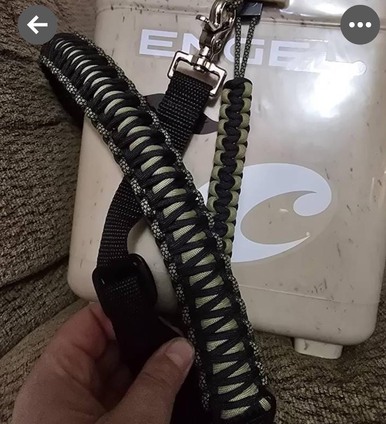 May include: Black and olive green paracord shoulder strap with a metal clasp and a matching wrist strap. The strap is attached to a beige cooler. The cooler has the brand name "ENGEL".