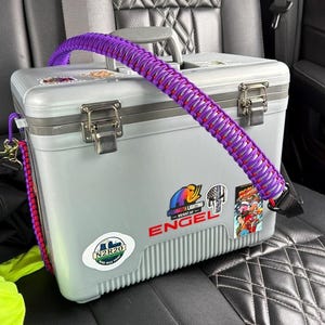 May include: A grey cooler with a purple and red braided strap. The cooler has metal latches and several stickers, including one that says "ENGEL". The cooler is on a black patterned car seat.