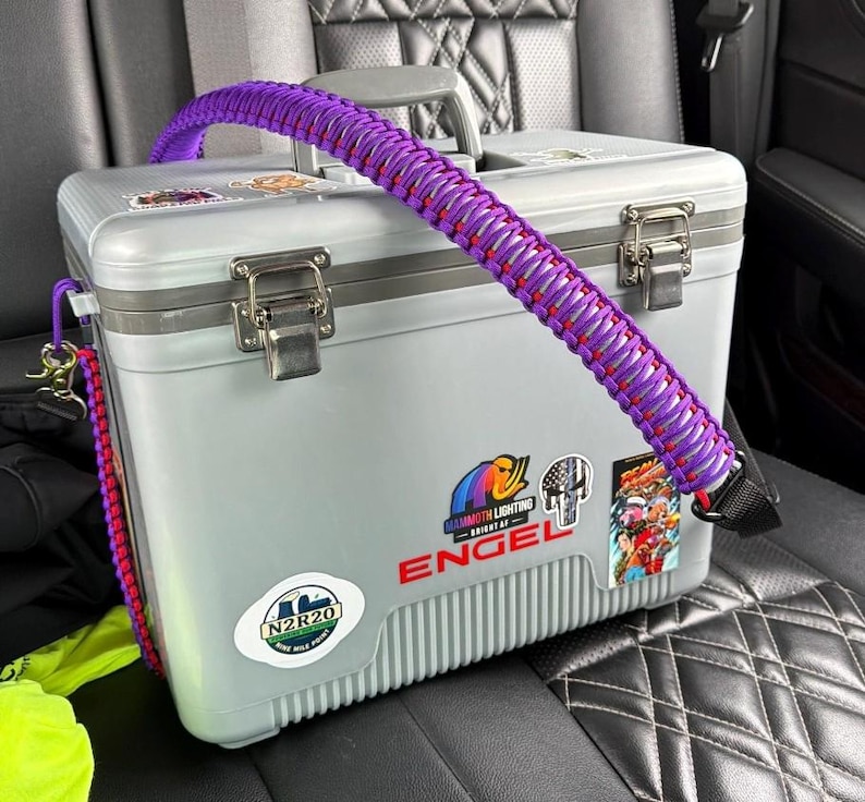 May include: A grey cooler with a purple and red braided strap. The cooler has metal latches and several stickers, including one that says "ENGEL". The cooler is on a black patterned car seat.