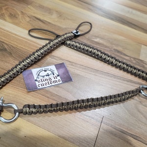 May include: A brown and beige paracord sling with a metal shackle and loop. The sling is made of woven cord and features the text "sling it customs" on a small card.