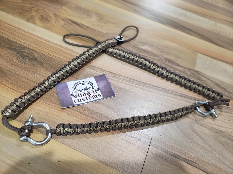 May include: A brown and beige paracord sling with a metal shackle and loop. The sling is made of woven cord and features the text "sling it customs" on a small card.