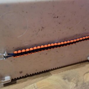 May include: A blue and orange paracord cooler strap with metal fixings. The strap is attached to a white cooler. The strap is made of woven nylon cord and is designed to secure the cooler lid.