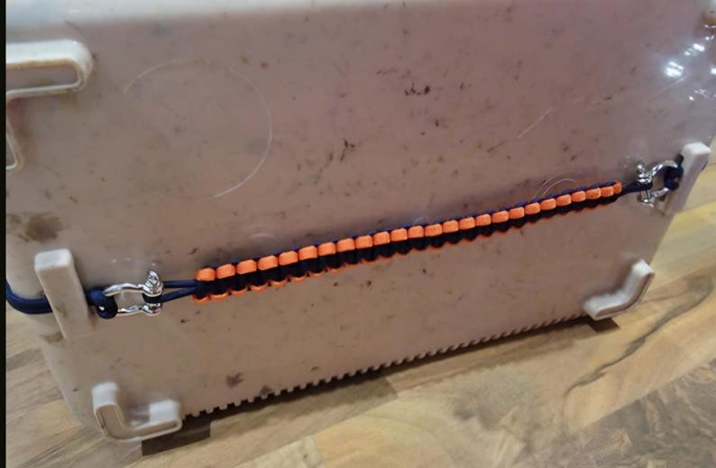 May include: A blue and orange paracord cooler strap with metal fixings. The strap is attached to a white cooler. The strap is made of woven nylon cord and is designed to secure the cooler lid.