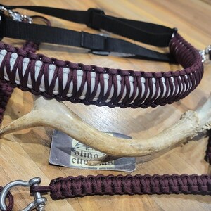 May include: A maroon and white paracord sling with a black adjustable strap and metal hardware. The sling is displayed with a deer antler and a small card. The text on the card is partially visible.