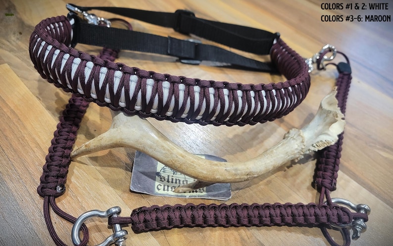 May include: A maroon and white paracord sling with a black adjustable strap and metal hardware. The sling is displayed with a deer antler and a small card. The text on the card is partially visible.