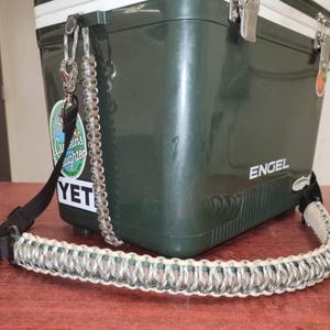 May include: A dark green cooler with a silver and white braided strap. The cooler has a black strap and a silver clasp. The word "ENGEL" is printed on the side. A sticker with the word "YETI" is visible.