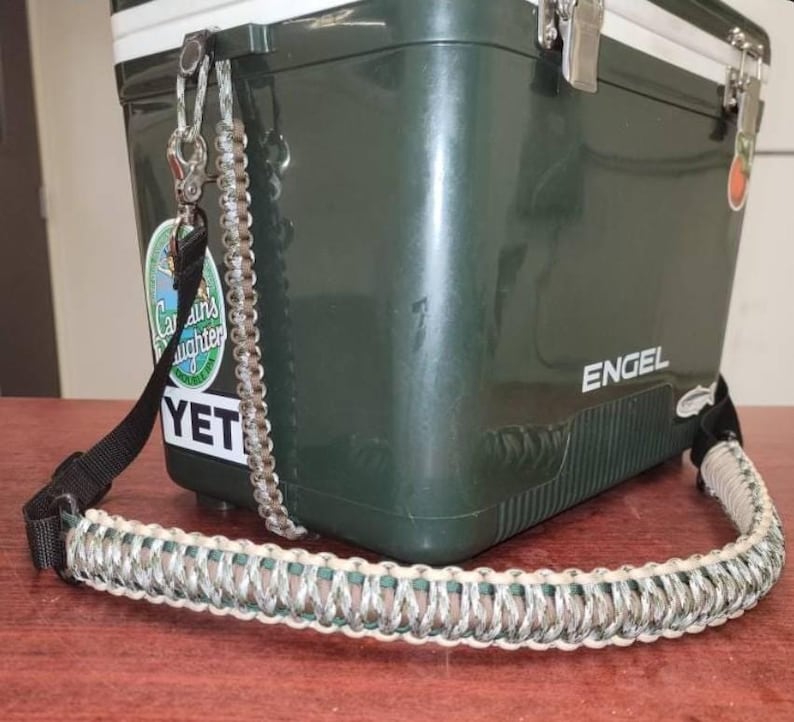 May include: A dark green cooler with a silver and white braided strap. The cooler has a black strap and a silver clasp. The word "ENGEL" is printed on the side. A sticker with the word "YETI" is visible.