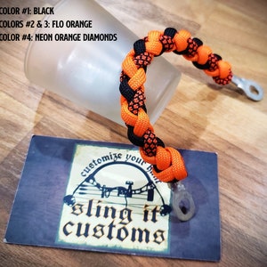 May include: Black and orange paracord bracelet with a metal loop. The bracelet is braided with a diamond pattern. The text "sling it customs" is visible on a card in the background.