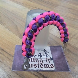 May include: A pink and purple paracord handle with metal loops. The handle is braided with a camouflage pattern. The handle is on a white surface with a business card that says "sling it customs" and "hunt".