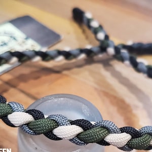 May include: A braided paracord bracelet with black, white, green, and gray strands. #1: BLACK #2: OLIVE GREEN