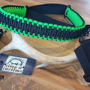 May include: A black and neon green paracord sling with two black straps. The sling is made with a braided paracord design. The text "sling it customs" is visible on a white card.