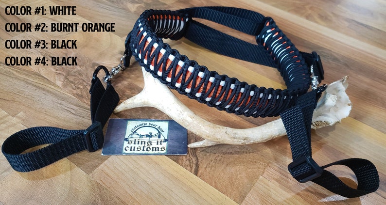 May include: A black and orange braided sling with white accents. The sling is made of paracord and has a black buckle and a silver swivel clip. The sling is displayed on a wooden surface with a white antler.