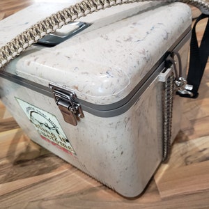 May include: A speckled, cream-coloured cooler with a braided handle and a black shoulder strap. The cooler has a metal latch and a sticker with text. A metal chain is attached to the side.