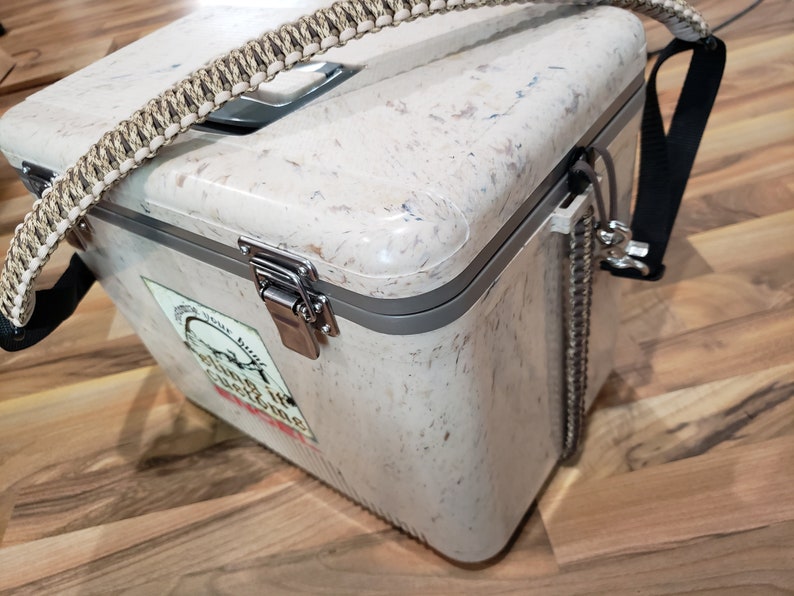 May include: A speckled, cream-coloured cooler with a braided handle and a black shoulder strap. The cooler has a metal latch and a sticker with text. A metal chain is attached to the side.