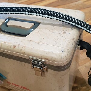 May include: A beige cooler with a black and white paracord handle. The cooler has two silver latches and a gray handle on the top. The cooler is sitting on a wooden surface.