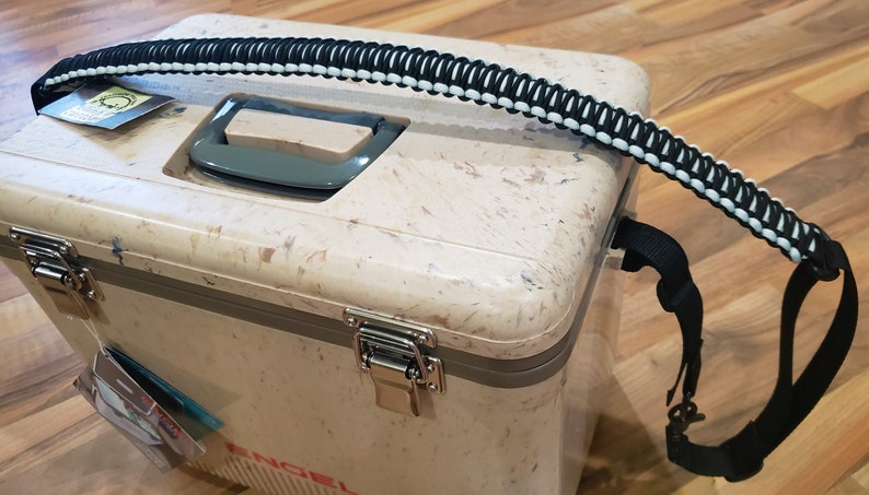May include: A beige cooler with a black and white paracord handle. The cooler has two silver latches and a gray handle on the top. The cooler is sitting on a wooden surface.