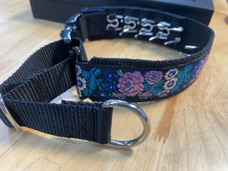 May include: A black nylon dog collar featuring a floral pattern with pink and blue flowers on the outer band. The collar includes a silver metal D-ring, a black plastic buckle, and a series of metal prongs lining the inner side of the strap.