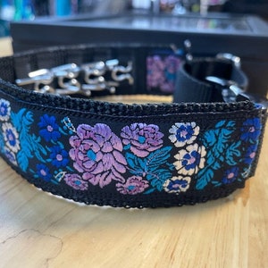 May include: A black collar featuring a vibrant floral embroidery pattern in shades of purple, light blue, and white. The collar is constructed from a durable black webbing material and includes a silver-toned metal prong buckle and hardware.