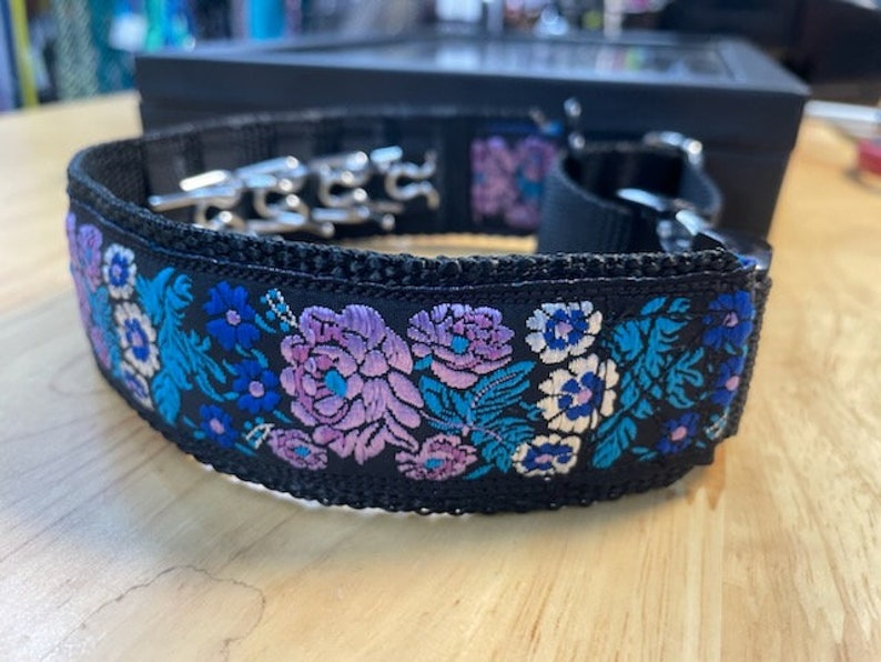 May include: A black collar featuring a vibrant floral embroidery pattern in shades of purple, light blue, and white. The collar is constructed from a durable black webbing material and includes a silver-toned metal prong buckle and hardware.