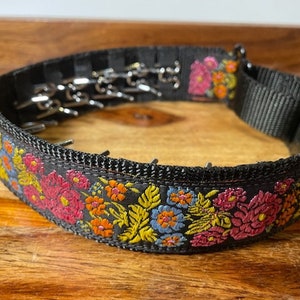 May include: A black nylon prong collar featuring a vibrant floral embroidery pattern in shades of pink, orange, yellow, and blue. The collar includes a series of inward-facing metal prongs, a black plastic side-release buckle, and a metal D-ring attachment.