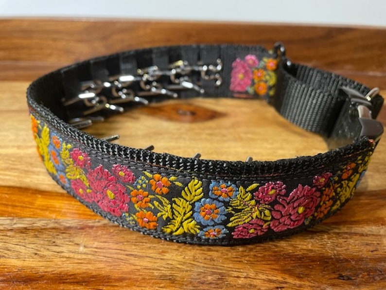 May include: A black nylon prong collar featuring a vibrant floral embroidery pattern in shades of pink, orange, yellow, and blue. The collar includes a series of inward-facing metal prongs, a black plastic side-release buckle, and a metal D-ring attachment.