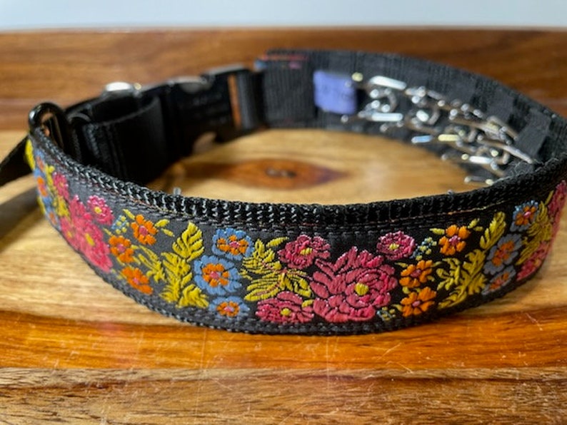 May include: A black nylon dog collar featuring a vibrant embroidered floral pattern in shades of pink, orange, yellow, and light blue. The collar includes a black plastic side-release buckle and a metal prong link chain section for adjustment.