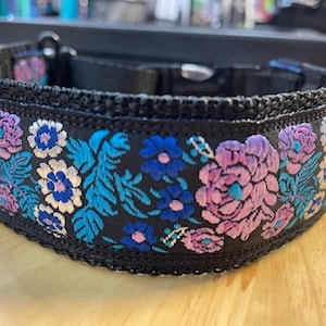 May include: A black pet collar featuring an intricate embroidered floral pattern in shades of vibrant blue, purple, and white. The collar is constructed with a black nylon webbing base, a black plastic side-release buckle, and a metal D-ring attachment.