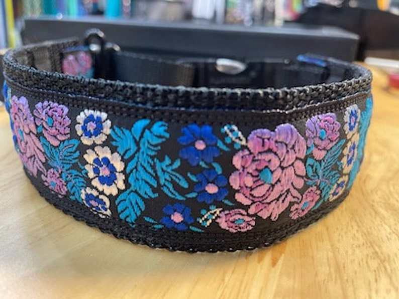 May include: A black pet collar featuring an intricate embroidered floral pattern in shades of vibrant blue, purple, and white. The collar is constructed with a black nylon webbing base, a black plastic side-release buckle, and a metal D-ring attachment.