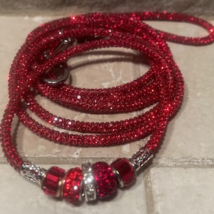 May include: A coiled red dog leash covered in sparkling crystals. The leash has a silver metal clasp and decorative beads at the handle end. The beads are red and silver, adding a touch of elegance to the pet accessory.