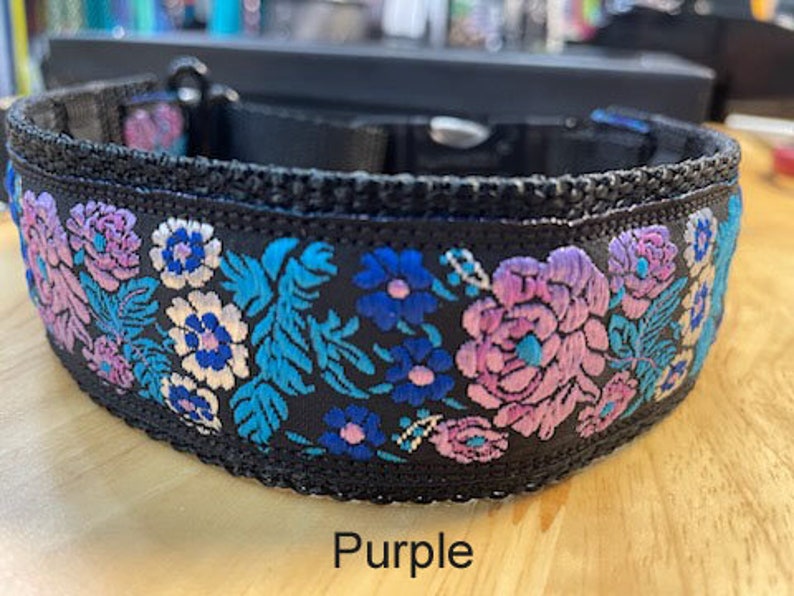 May include: A black pet collar featuring an embroidered floral pattern in shades of purple, light blue, and white. The collar includes a black plastic side-release buckle and a metal D-ring. The word Purple is printed below the collar.