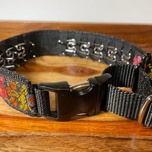 May include: A black nylon pet collar featuring a floral pattern with red, orange, and blue flowers on one side. The collar includes a black plastic side-release buckle, a metal adjustment slider, and a series of silver-toned metal prongs along the inner band.