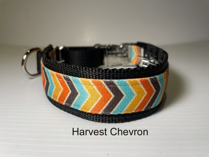 May include: A black dog collar with a colourful chevron pattern in orange, yellow, blue, and grey. The collar has a silver buckle and a D-ring. The text "Harvest Chevron" is printed below the collar.
