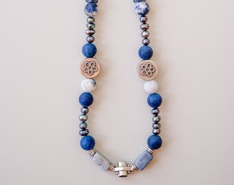 May include: A blue and white beaded necklace with silver accents. The necklace features round and rectangular beads, as well as two silver flower-shaped charms.