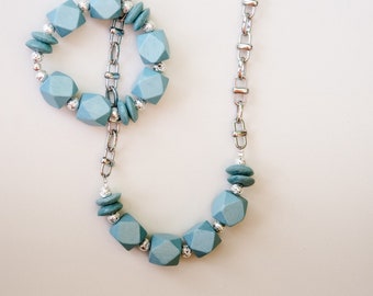 May include: A silver chain necklace with a light blue wooden bead pendant. The pendant is a circle of geometrically shaped beads.