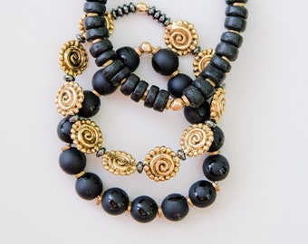 May include: Three black beaded bracelets with gold accents. The bracelets are stacked on top of each other, with the largest bracelet on the bottom and the smallest bracelet on top. The gold accents are in the shape of swirls and circles.