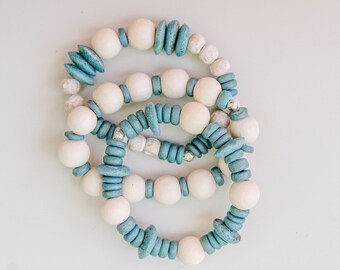 May include: Three stacked bracelets made of white and blue wooden beads. The bracelets are made of round beads and flat, oval-shaped beads.