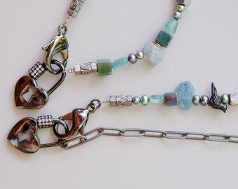 May include: A silver chain necklace with a heart-shaped clasp and a variety of blue and green beads. The necklace is made of metal and has a delicate, feminine look.