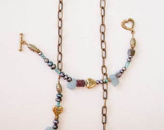 May include: A gold chain necklace with a heart-shaped clasp and a variety of beads, including turquoise, purple, and gold. The necklace is hanging from a silver clipboard.