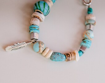 May include: A turquoise and white beaded bracelet with a silver feather charm. The bracelet features a variety of turquoise and white beads, including some with a marbled pattern.