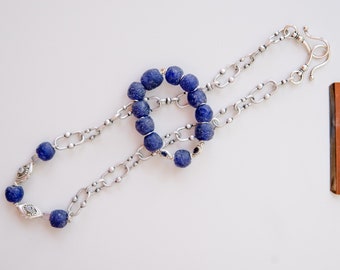 May include: A necklace featuring dark blue, round beads and a silver chain. The chain has a series of oval links and decorative elements. The necklace is displayed on a white surface. A brown and silver object is in the background.