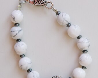 May include: A white and silver bracelet with a heart-shaped clasp and a large silver bead with an intricate design. The bracelet is made of white beads with black accents.