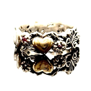 Puede incluir: Un anillo de plata con un corazón dorado y un diseño floral. El anillo presenta pequeñas piedras preciosas rojas y detalles intrincados. El anillo está sobre un fondo blanco, resaltando sus características ornamentadas.