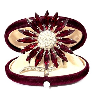 Peut inclure: Broche florale vintage avec un centre de cristaux clairs entouré de pierres rouges rubis taillées en marquise. La broche est sertie dans un métal argenté et présentée dans un écrin en velours bordeaux avec un intérieur crème.