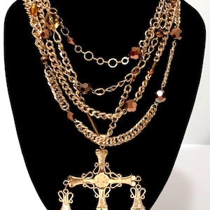 May include: A multi-layered gold-tone necklace with a large cross pendant. The necklace features various chain styles, including curb and beaded chains, and is adorned with brown and amber-colored beads. The cross pendant has ornate detailing and bell-shaped dangles.