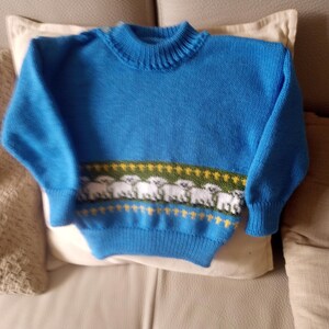 Pode incluir: Um suéter azul de malha com gola redonda e mangas compridas. O suéter apresenta uma faixa horizontal com um padrão de ovelhas brancas sobre um fundo verde, acentuado com detalhes amarelos. O suéter é provavelmente para uma criança.