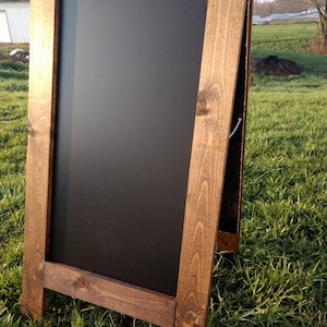 May include: A brown wooden A-frame chalkboard sign with a black chalkboard surface. The sign is standing on green grass.