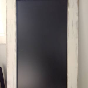 May include: A whitewashed wooden frame surrounds a black chalkboard. The frame has a distressed look with visible wood grain and white paint.