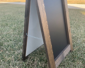 May include: A wooden A-frame chalkboard sign with a dark brown stain. The sign has a chain attached to the top to help keep it closed.